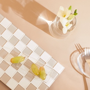 May include: A white and beige tiled surface with a clear glass vase filled with white flowers. A clear glass bowl with a silver fork sits on a beige surface. A clear glass container with green grapes sits on the tiled surface.