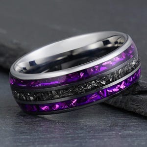 Peut inclure: Une bague en métal argenté présentant une bande centrale de matériau gris foncé texturé, encadrée par deux sections incrustées de couleur violet vif. La bague possède une finition polie et réfléchissante sur les bords et un profil arrondi.