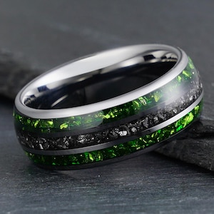 Puede incluir: Un anillo de metal plateado pulido con una incrustación central de material negro oscuro y texturizado, flanqueado por dos bandas de piedra triturada de color verde vibrante y brillante que rodean todo el anillo.