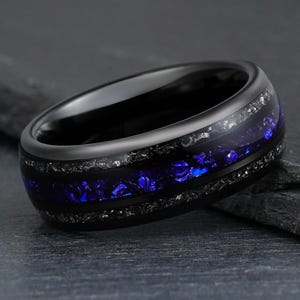 Op de afbeelding: Een zwarte ring met een centrale inleg van levendig blauw verbrijzeld materiaal, geflankeerd door twee dunne banden van glinsterende zilverkleurige spikkels. De ring heeft een gladde, gepolijste afwerking en een afgerond profiel met diep contrast.
