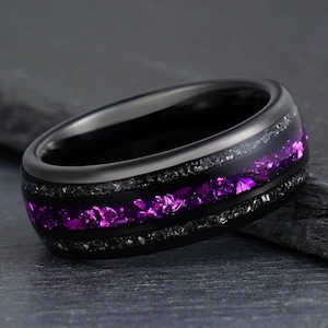 May include: A black ring featuring a central inlay of vibrant purple crushed material flanked by two thin bands of dark, shimmering, granular texture. The band has a smooth, polished finish and a rounded profile, creating a distinct contrast.