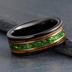 May include: A black ring featuring a central inlay of crushed green stone fragments flanked by two thin metallic bands. The outer edges of the ring are smooth and dark, creating a contrast with the vibrant green texture and the warm metallic tones.