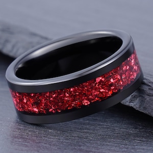 May include: A black ring featuring a wide, recessed centre channel filled with crushed red stone inlay. The band has a smooth, polished finish on the edges, contrasting with the textured, vibrant red material embedded within the dark metal surface.