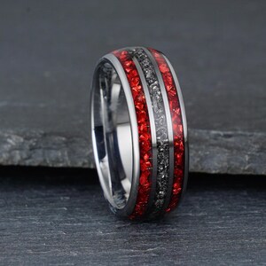 Peut inclure: Une bague en m&eacute;tal argent&eacute; dot&eacute;e d'une incrustation centrale en mat&eacute;riau gris fonc&eacute; textur&eacute;, entour&eacute;e de deux incrustations en pierre rouge broy&eacute;e. L'anneau pr&eacute;sente une finition polie et r&eacute;fl&eacute;chissante sur les bords et un profil arrondi.