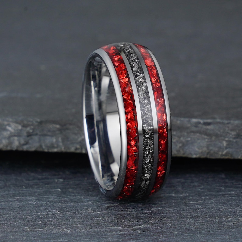 Peut inclure: Une bague en m&eacute;tal argent&eacute; dot&eacute;e d'une incrustation centrale en mat&eacute;riau gris fonc&eacute; textur&eacute;, entour&eacute;e de deux incrustations en pierre rouge broy&eacute;e. L'anneau pr&eacute;sente une finition polie et r&eacute;fl&eacute;chissante sur les bords et un profil arrondi.