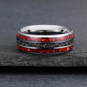 Peut inclure: Une bague anneau en m&eacute;tal argent&eacute; poli avec une incrustation centrale de mat&eacute;riau gris fonc&eacute; textur&eacute;, entour&eacute;e de deux bandes rouges vibrantes rappelant de la pierre concass&eacute;e. Les bords pr&eacute;sentent une finition m&eacute;tallique lisse.