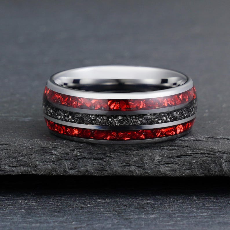 Peut inclure: Une bague anneau en m&eacute;tal argent&eacute; poli avec une incrustation centrale de mat&eacute;riau gris fonc&eacute; textur&eacute;, entour&eacute;e de deux bandes rouges vibrantes rappelant de la pierre concass&eacute;e. Les bords pr&eacute;sentent une finition m&eacute;tallique lisse.