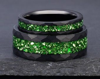 Könnte beinhalten: Zwei schwarze Ringe mit gehämmertem Finish und einer zentralen Einlage aus zerkleinertem grünem Stein. Die Ringe haben eine dunkle, matte Außenstruktur, die einen Kontrast zu den hellen, reflektierenden und gezackten grünen Mineralpartikeln bildet.