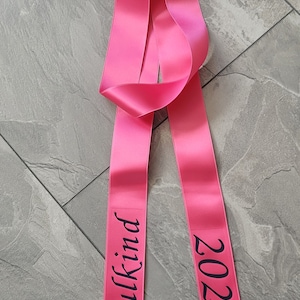 Könnte beinhalten: Ein rosa Satinband mit schwarzem Text, der "Schulkid 2024" lautet.