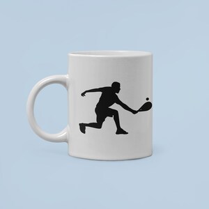 May include: White ceramic mug with a black silhouette of a person playing paddleball. The person is in mid-swing, holding a paddle and a ball is in the air.