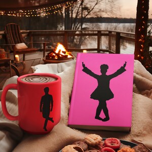 May include: A red mug with a silhouette of a person dancing on it, a pink book with a silhouette of a person dancing on the cover, and a plate of cookies on a brown surface.