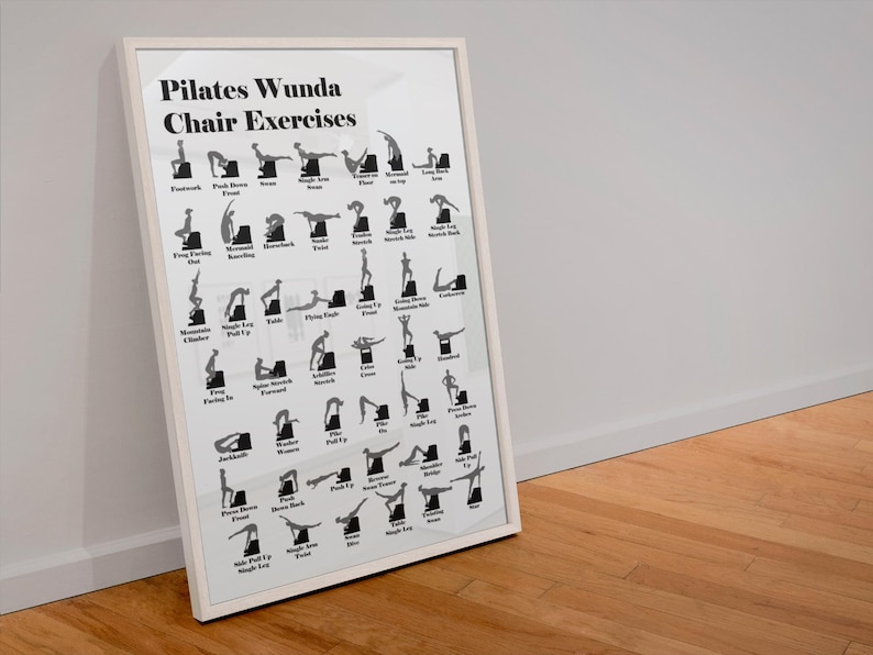May include: A framed Pilates Wunda Chair Exercises poster. The white poster features black silhouettes demonstrating various exercises. The poster is in a white frame and leans against a white wall and a wooden floor.