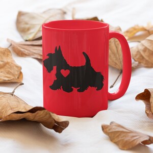 May include: A red ceramic mug with a black silhouette of a Scottish Terrier with a red heart on its chest. The mug is surrounded by brown leaves on a white background.