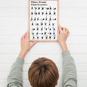 May include: A framed Pilates Wunda Chair Exercises poster. The poster features black and white illustrations of various exercises. The frame is a light brown color. The poster is being held up against a white brick wall.