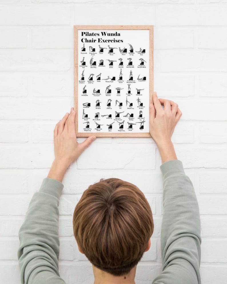 May include: A framed Pilates Wunda Chair Exercises poster. The poster features black and white illustrations of various exercises. The frame is a light brown color. The poster is being held up against a white brick wall.
