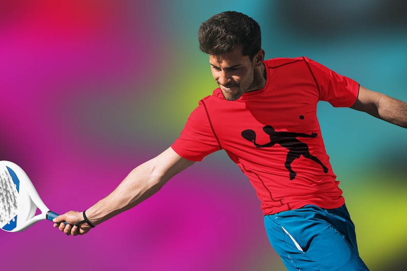 May include: A man wearing a red t-shirt with a black silhouette of a person playing paddle tennis. He is holding a paddle tennis racket and is in mid-swing.
