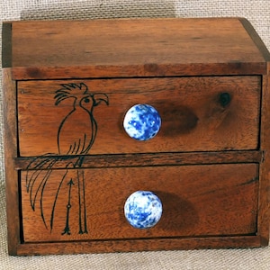 May include: A small wooden chest with two drawers. The drawers have blue and white ceramic knobs. The front of the chest is decorated with a black and white drawing of a bird.