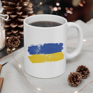 Puede incluir: Taza de cerámica blanca con un diseño de pinceladas azules y amarillas que se asemeja a la bandera de Ucrania. La taza está llena de café y se encuentra sobre una superficie de tela gris con luces de cadena.