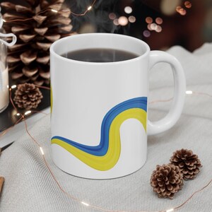 Puede incluir: Taza de cerámica blanca con un diseño abstracto azul y amarillo. La taza está llena de café y tiene un asa.