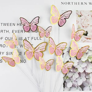 May include: A selection of pink and gold butterfly decorations on clear stems. The butterflies feature a pink body with gold edging, arranged in a decorative display. The background includes text and floral elements.