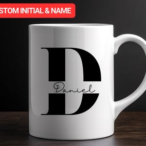 Può includere: Tazza in ceramica bianca con una grande lettera "D" nera e il nome "Daniel" in corsivo. La tazza ha un manico curvo e il testo "CUSTOM INITIAL & NAME" in un banner rosso nella parte superiore.