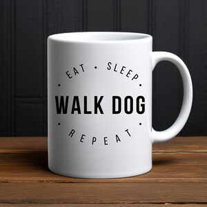 Könnte beinhalten: Weiße Keramik-Tasse mit dem schwarzen Text "EAT SLEEP WALK DOG REPEAT" in kreisförmiger Anordnung. Die Tasse hat einen Henkel und steht auf einer Holzoberfläche.
