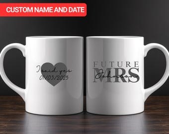 Op de afbeelding: Twee witte keramische mokken. Eén mok heeft de tekst "I said yes" met een hart en de datum 01/03/2025. De andere mok zegt "FUTURE MRS Johnson". De mokken staan op een donker houten oppervlak.