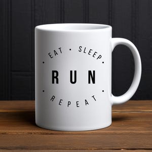 Könnte beinhalten: Weiße Keramik-Kaffeetasse mit schwarzem Text, der "Essen. Schlafen. Laufen. Wiederholen." lautet.