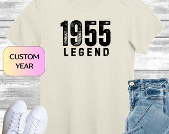 Może przedstawiać: Kremowa koszulka z napisem "1955 LEGEND" pogrubioną, postarzaną czarną czcionką. Niestandardowa etykieta z rokiem znajduje się w lewym dolnym rogu. Koszulka jest zestawiona z niebieskimi dżinsami i białymi trampkami, sugerując styl casual.