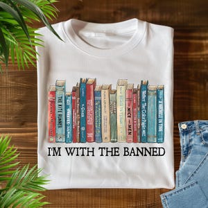 Könnte beinhalten: Weißes T-Shirt mit einer Grafik eines Bücherregals mit Buchtiteln wie "The Catcher in the Rye" und "The Handmaid's Tale". Der Text "I'M WITH THE BANNED" ist darunter gedruckt. Das Shirt besteht aus weichem Material.