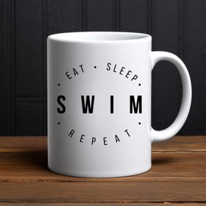 May include: White ceramic coffee mug with black text that reads "Eat. Sleep. Swim. Repeat."