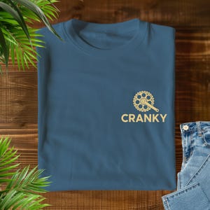 Peut inclure: Un t-shirt bleu sarcelle avec un motif de pédalier de vélo doré et le mot "CRANKY" imprimé en dessous. Le t-shirt est plié sur une surface en bois, avec un jean bleu et des feuilles vertes en arrière-plan.
