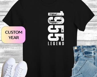 Może przedstawiać: Czarny t-shirt z białym napisem "1955 LEGEND" w pionowej czcionce. Obraz zawiera również parę białych tenisówek, niebieskie dżinsy i etykietę "CUSTOM YEAR".