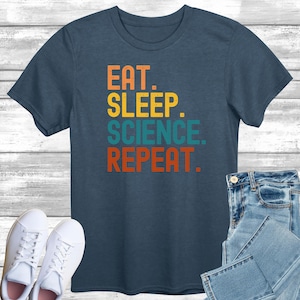 Puede incluir: Una camiseta azul marino con las palabras "EAT. SLEEP. SCIENCE. REPEAT." en letras de bloque naranja, amarillo, turquesa y rojo. La camiseta se combina con vaqueros azules y zapatillas blancas.