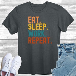May include: Dark gray t-shirt with the text "EAT. SLEEP. WORK. REPEAT." in orange, yellow, teal, and red. The shirt is paired with blue jeans and white sneakers.