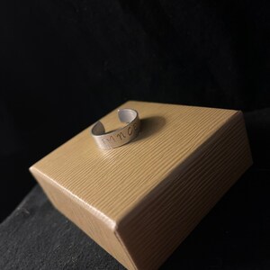 Op de afbeelding: Een zilverkleurige verstelbare ring met het woord "DANCE" aan de buitenkant gestempeld. De ring ligt op een kleine, bruinachtige, getextureerde doos tegen een donkere achtergrond.