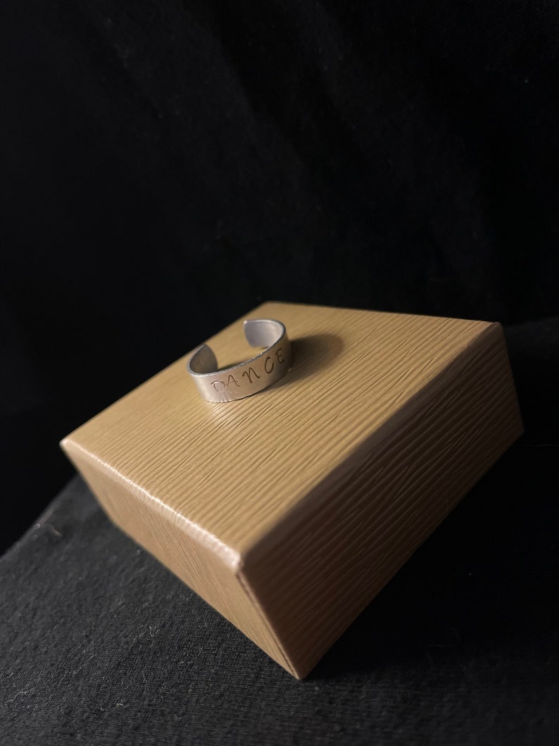 Op de afbeelding: Een zilverkleurige verstelbare ring met het woord "DANCE" aan de buitenkant gestempeld. De ring ligt op een kleine, bruinachtige, getextureerde doos tegen een donkere achtergrond.