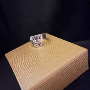 K&ouml;nnte beinhalten: Ein silberner Wickelring mit den Buchstaben "GTR" darauf gestempelt. Der Ring wird auf einer strukturierten, hellbraunen Oberfl&auml;che vor dunklem Hintergrund pr&auml;sentiert. Die Buchstaben sind in handschriftlichem Stil.