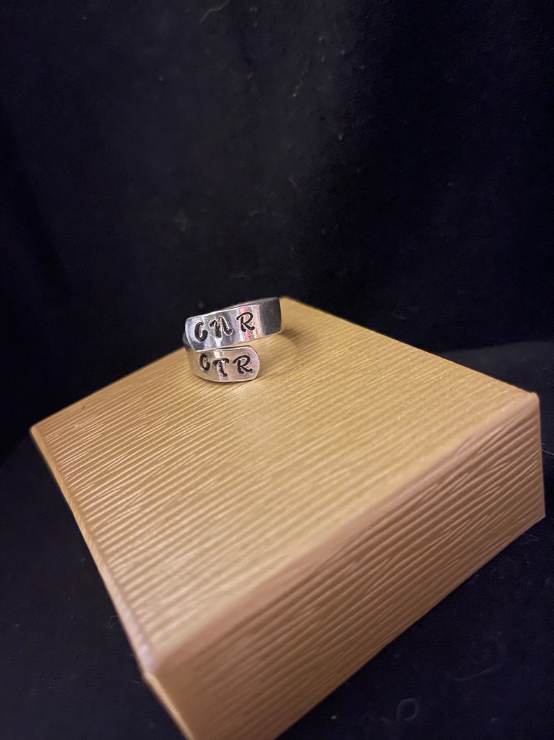 K&ouml;nnte beinhalten: Ein silberner Wickelring mit den Buchstaben "GTR" darauf gestempelt. Der Ring wird auf einer strukturierten, hellbraunen Oberfl&auml;che vor dunklem Hintergrund pr&auml;sentiert. Die Buchstaben sind in handschriftlichem Stil.