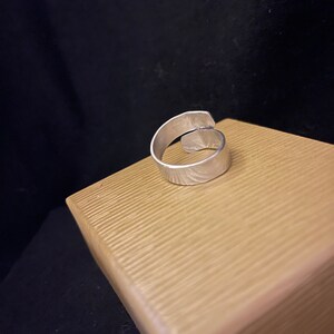 K&ouml;nnte beinhalten: Ein silberfarbener, verstellbarer Ring mit strukturierter Oberfl&auml;che. Der Ring liegt auf einer hellbraunen, strukturierten Oberfl&auml;che vor dunklem Hintergrund. Das Design des Rings weist ein offenes, &uuml;berlappendes Band auf.