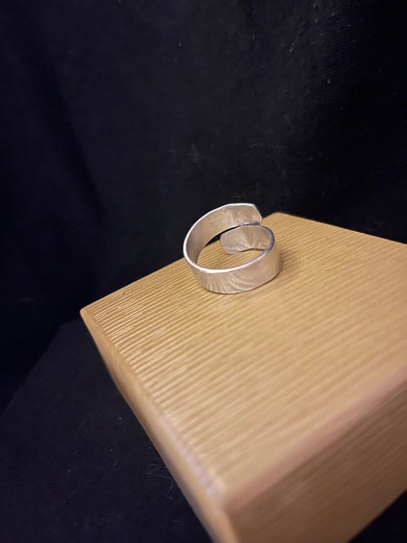 K&ouml;nnte beinhalten: Ein silberfarbener, verstellbarer Ring mit strukturierter Oberfl&auml;che. Der Ring liegt auf einer hellbraunen, strukturierten Oberfl&auml;che vor dunklem Hintergrund. Das Design des Rings weist ein offenes, &uuml;berlappendes Band auf.