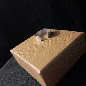 Op de afbeelding: Een zilverkleurige, open ring. De ring ligt op een lichtbruine, getextureerde doos. De achtergrond is een donkere, effen kleur. De ring lijkt van metaal te zijn en heeft een eenvoudig, minimalistisch ontwerp.