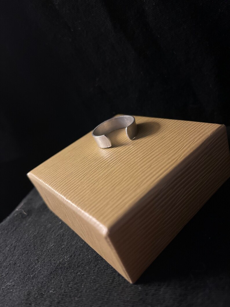 Op de afbeelding: Een zilverkleurige, open ring. De ring ligt op een lichtbruine, getextureerde doos. De achtergrond is een donkere, effen kleur. De ring lijkt van metaal te zijn en heeft een eenvoudig, minimalistisch ontwerp.