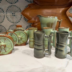 Pode incluir: Uma coleção de cerâmica artesanal em tons de verde e castanho. O conjunto inclui uma chaleira, jarras e canecas, todas com um design rústico em forma de roda. A cerâmica é exibida numa bancada branca com um painel de fundo estampado.