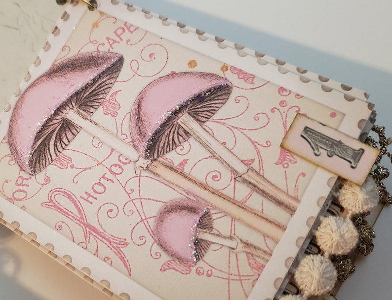 May include: A pink and white scrapbook with a decorative mushroom design and the word "Hotdog" printed on the cover. The scrapbook has a white tassel trim and a pink tag with the number "1" on it.