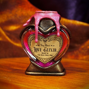 Puede incluir: Una botella de vidrio en forma de corazón llena de líquido rosa, etiquetada como "Love Elixir". La botella tiene un marco de metal color bronce y un sello de cera rosa. También es visible el texto "Very Potent" y "Brewed by The Secret Moon Shop".