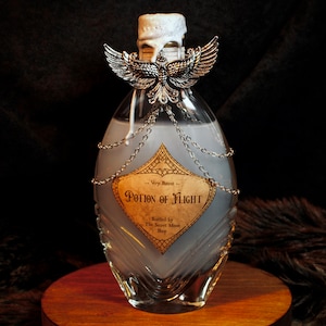 Könnte beinhalten: Eine klare Glas-Zaubertrankflasche mit silberner Vogelverzierung und dem Etikett "Potion of Flight". Die Flasche ist mit einer hellblauen Flüssigkeit gefüllt und mit weißem Wachs versiegelt. Dekorative Ketten schmücken die Flasche.