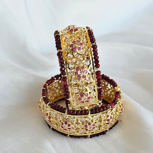 May include: Two gold bangles with intricate designs, adorned with small pink and white gemstones. The bangles are surrounded by a string of dark red beads.