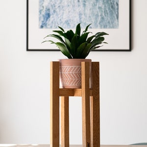 May include: A wooden plant stand with four legs, supporting a pink planter with a white chevron pattern. A green plant with large leaves is in the planter. The stand is on a light wood surface.