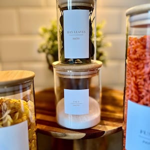 Può includere: Barattoli di vetro trasparente con coperchi in bambù, etichettati "BAY LEAVES" e "SALT" in una dispensa. I barattoli contengono foglie di alloro e sale. Altri barattoli contengono alimenti. I barattoli sono su un vassoio di legno.